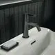 Waschtischarmatur in Magnet Grau von Ideal Standard, montiert auf einer weißen Oberfläche, neben einem Seifenhalter. Hintergrund mit schwarzen Fliesen.